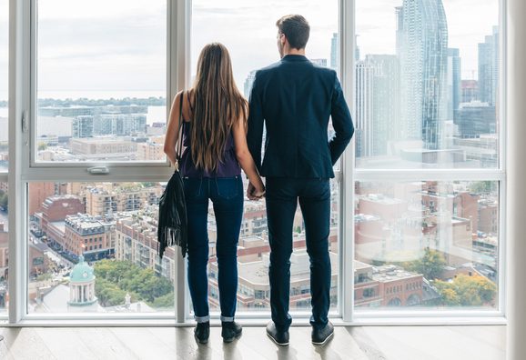 A young couple holding hands as they look out the window of a condominium unit. They are shopping for real estate in the city.